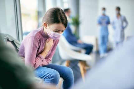 Девчока в медицинской маске сидит в коридоре больницы и держится за сердце.