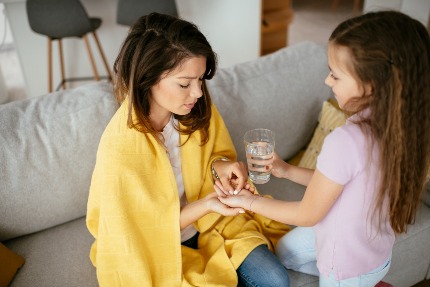 Маленькая девочка протягивает заболевшей маме таблетку и стакан с водой.