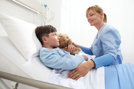 Мальчик лежит на больничной кровати с мягкой игрушкой, рядом сидит мама.