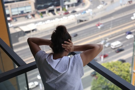 Девушка стоит на балконе высокого здания, закрыв уши руками.