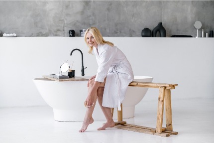 Женщина делает массаж сухой щеткой, сидя на скамеечке у ванны.