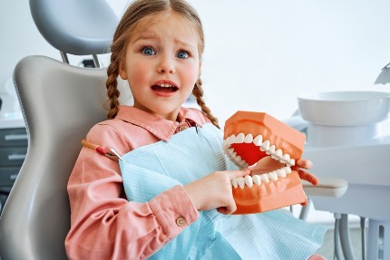 Девочка сидит в стоматологическом кресле, в руках держит макет челюстей.