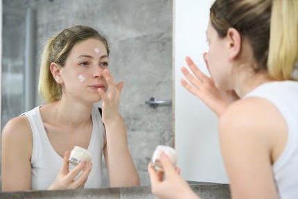 Женщина наносит крем на лицо у зеркала.