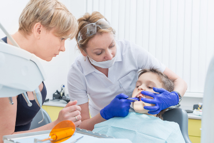 Стоматолог лечит зуб мальчику, рядом стоит его мама.