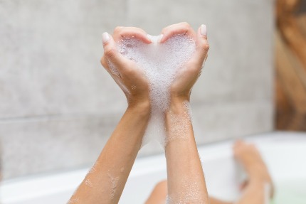 Женщина, которая принимает ванну, сложила руки с пеной в виде сердечка.