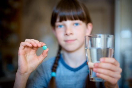Девочка с косичками держит в одной руке таблетку, а в другой стакан воды
