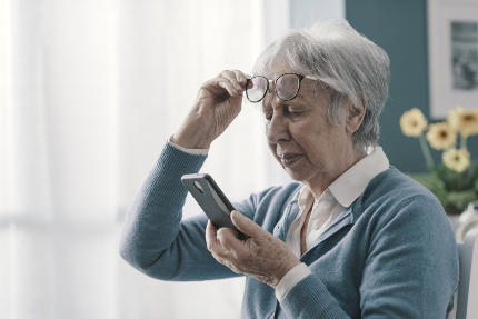 Пожилая женщина надевает очки, чтобы прочесть что-то в телефоне.