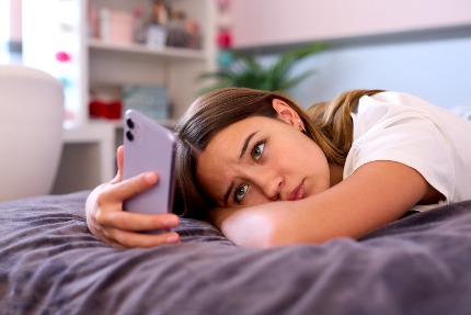 Грустная девушка лежит на кровати и смотрит на телефон.